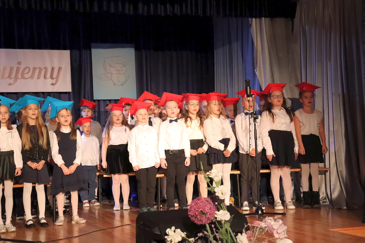 Uczniowie klas pierwszych śpiewają piosenkę “Szkoło, szkoło…” ubrani w birety niebieskie i czerwone na tle napisu Ślubujemy