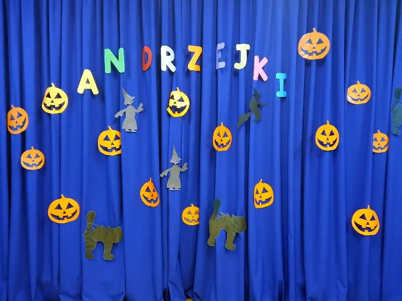 Na niebieskim tle napis „Andrzejki” i elementy dekoracyjne: dynie, czarownice, koty.