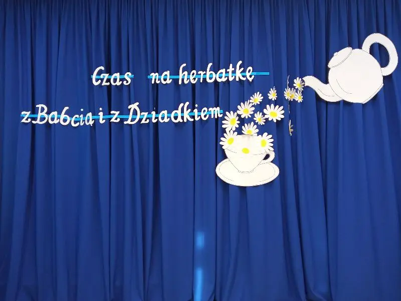fotografia dekoracji - na niebieskiej kotarze biały napis: Czas na herbatkę z Babcią i Dziadkiem, rysunek: z dzbanka przelewają się kwiaty do filiżanki