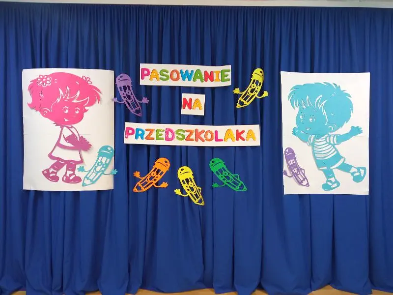 Dekoracja małej auli: napis „Pasowanie na Przedszkolaka” otoczony kolorowymi kredkami, po prawej stronie - postać chłopca a po lewej dziewczynki.