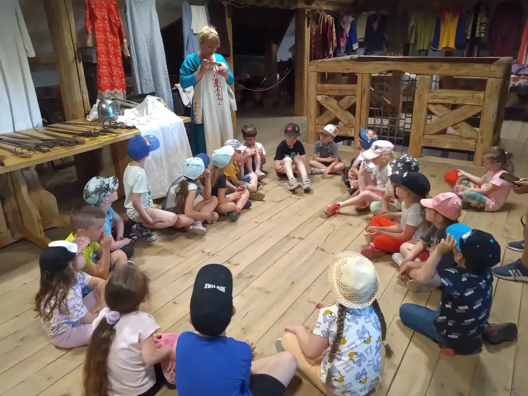 Dzieci siedzą w kole na podłodze w pomieszczeniu zamkowym zwanym garderobą i prowadząca omawia i prezentuje suknię lnianą