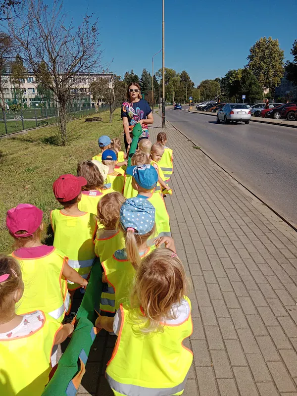 Dzieci trzymają się węża spacerowego i idą chodnikiem wzdłuż jezdni po chodniku