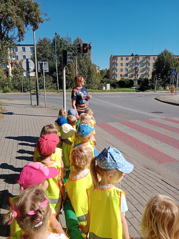 Dzieci stoją w parach na chodniku przed przejściem na skrzyżowaniu z sygnalizacją świetlną