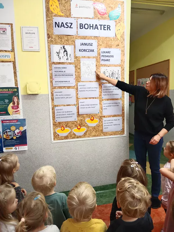 Dzieci zgromadzone przed tablicą w hollu przedszkola upamiętniającą Janusza Korczaka